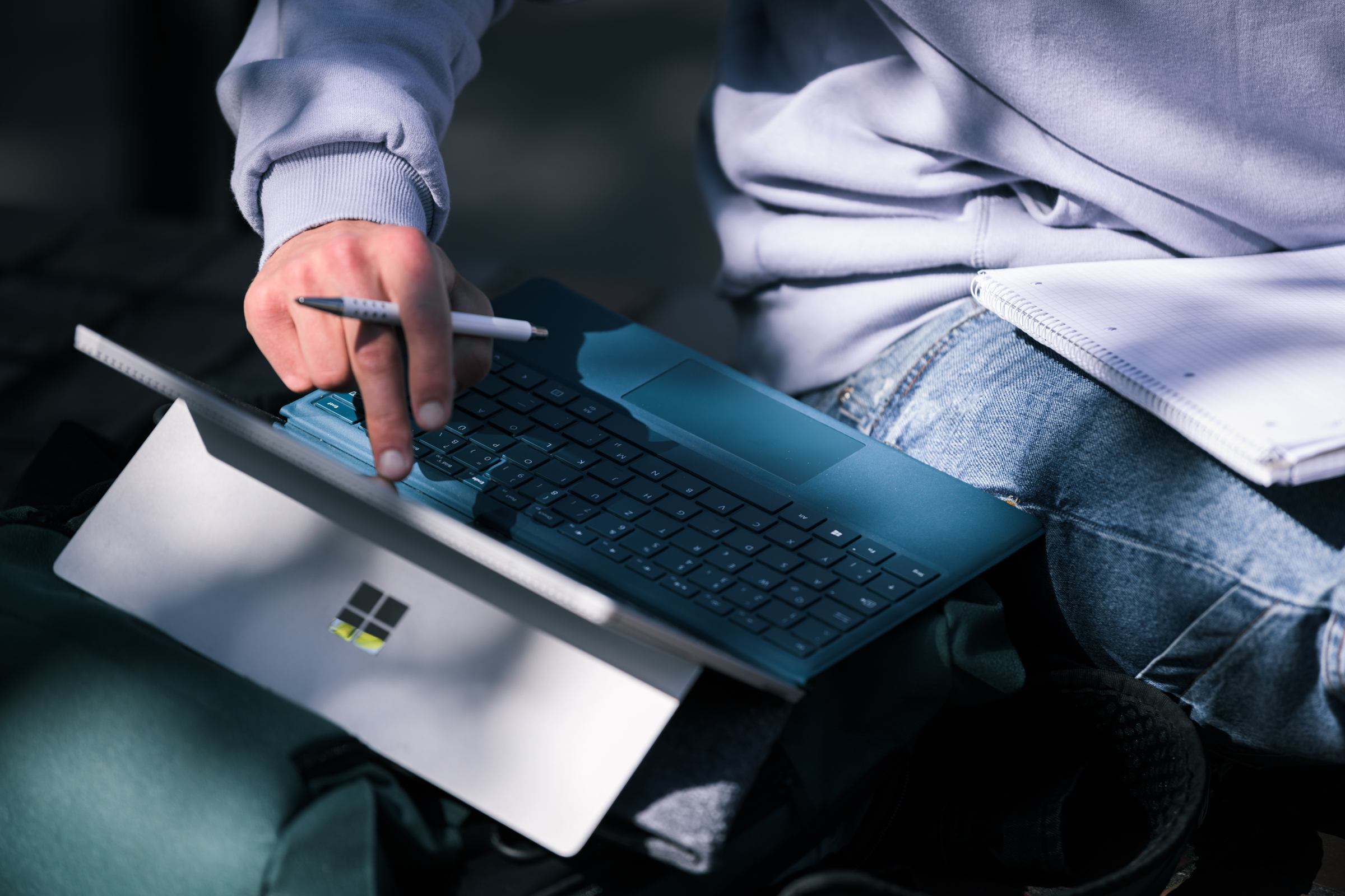 Zu sehen ist eine Hand mit einem zwischen Zeige- und Mittelfinger eingeklemmten Stift, die auf den Bildschirm eines Laptops deutet.